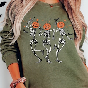 May include: Green long-sleeved sweatshirt with a graphic of three skeletons wearing pumpkin heads. The skeletons are dancing and have a fun, spooky vibe.