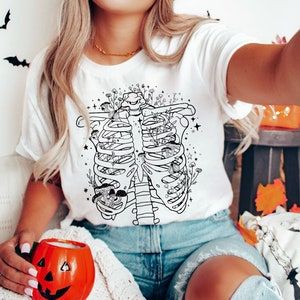 May include: A white t-shirt with a black line drawing of a ribcage filled with flowers and mushrooms. The shirt is being worn by a person with blonde hair and blue jeans.