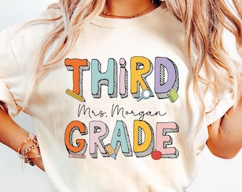 May include: Cream-colored t-shirt with the words "THIRD GRADE" in colorful, outlined letters. The text "Mrs. Morgan" is written below the word "THIRD". The shirt is paired with light blue denim jeans.