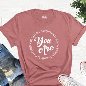 May include: A dusty rose t-shirt with a white circle graphic that says "You Are" surrounded by the words "worthy, important, kind, strong, capable, loved".