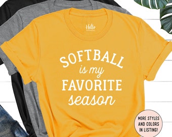 May include: Yellow t-shirt with the text "Softball is my favorite season" in white lettering. The shirt is folded and displayed on a white surface with a pair of jeans and other shirts.
