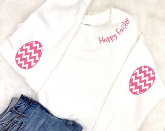 Könnte beinhalten: Ein weißes Sweatshirt mit dem Schriftzug "Happy Easter" in rosa. Die Ärmel zeigen rosa Osterei-Designs mit einem weißen Zickzackmuster. Blaue Jeans sind neben dem Sweatshirt gefaltet.