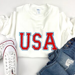 May include: A white long-sleeve crewneck sweatshirt featuring the letters USA in red with a blue glitter outline. The sweatshirt is paired with blue denim jeans that have a distressed knee detail and a pair of white canvas sneakers with red trim.
