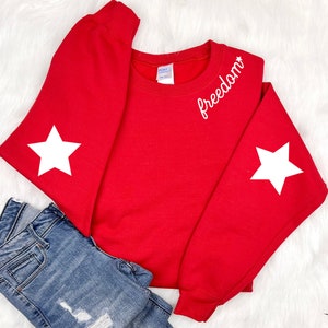 May include: Red sweatshirt with white stars on the sleeves and the word "freedom" embroidered on the front.