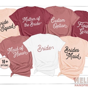 May include: A collection of seven short-sleeved crew neck t-shirts in shades of white, light pink, and maroon. The shirts feature white or black cursive text including Bride Squad, Mother of the Bride, Custom Option, Flower Girl, Maid of Honor, Bride, and Brides Maid.