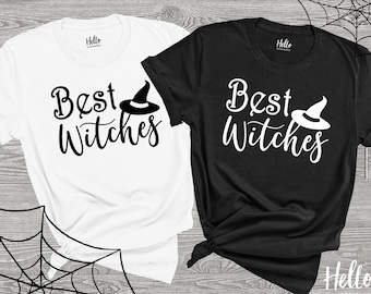 May include: Two black and white t-shirts with the text "Best Witches" and a witch's hat graphic. The shirts are laid out on a wooden surface with a spiderweb in the background.