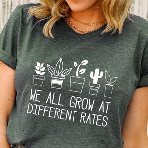 May include: A heather forest green short-sleeve t-shirt featuring a white graphic of five potted plants in a row above the text "WE ALL GROW AT DIFFERENT RATES" in a clean, sans-serif font. The shirt has a crew neckline and cuffed sleeves.