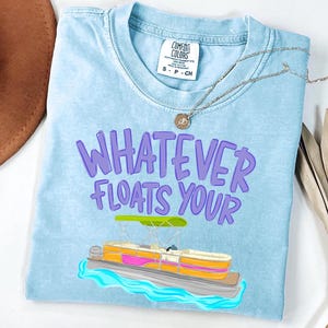 Puede incluir: Camiseta azul claro con la frase "WHATEVER FLOATS YOUR" en letras moradas. Debajo, una colorida ilustraci&oacute;n de una lancha pont&oacute;n. Un collar plateado con un colgante est&aacute; sobre la camiseta.