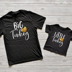 May include: Two black t-shirts with a gold turkey graphic and white text. The larger shirt says "BIG TURKEY" and the smaller shirt says "LITTLE TURKEY".