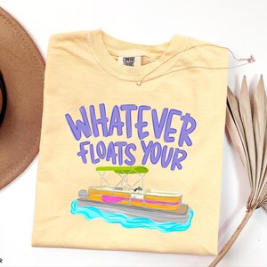 Puede incluir: Camiseta de color mantequilla con la frase "WHATEVER FLOATS YOUR" en letras moradas. Una colorida ilustraci&oacute;n de un barco pont&oacute;n est&aacute; debajo del texto. La camiseta es de la marca Comfort Colors.