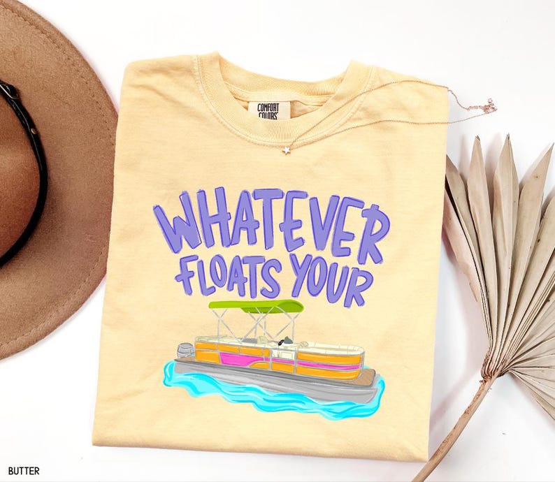 Puede incluir: Camiseta de color mantequilla con la frase "WHATEVER FLOATS YOUR" en letras moradas. Una colorida ilustraci&oacute;n de un barco pont&oacute;n est&aacute; debajo del texto. La camiseta es de la marca Comfort Colors.