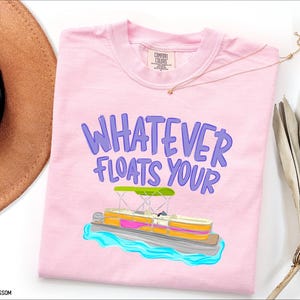 Puede incluir: Camiseta rosa claro con la frase "WHATEVER FLOATS YOUR" en morado. Debajo, una colorida ilustraci&oacute;n de un barco pont&oacute;n con toldo verde y agua azul.