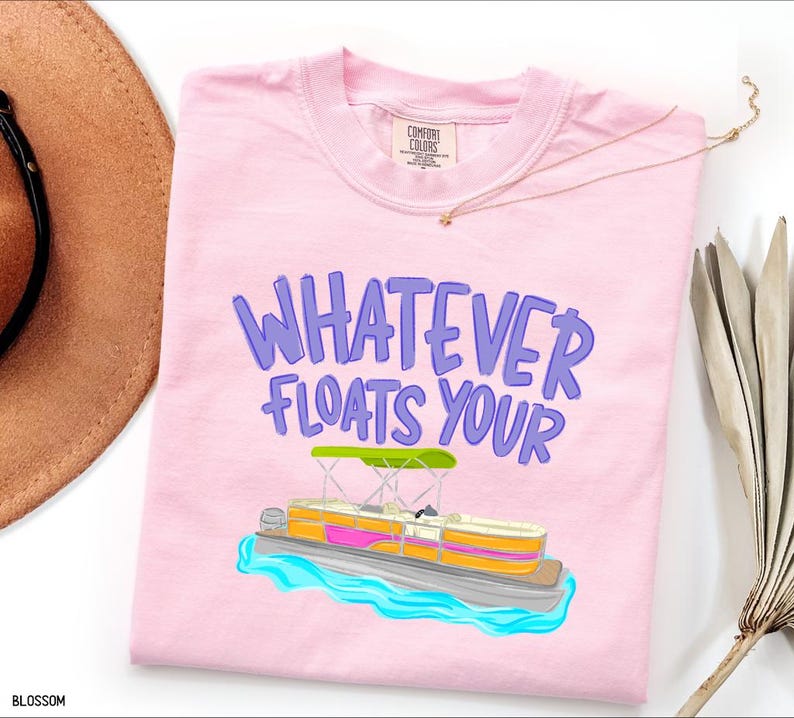 Puede incluir: Camiseta rosa claro con la frase "WHATEVER FLOATS YOUR" en morado. Debajo, una colorida ilustraci&oacute;n de un barco pont&oacute;n con toldo verde y agua azul.