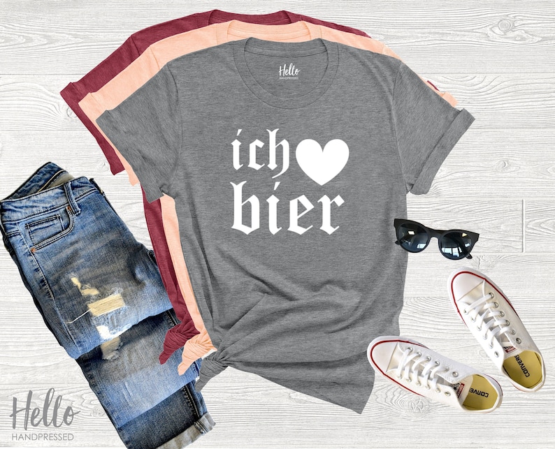 Op de afbeelding: Een grijs T-shirt met de tekst "ich &hearts; bier" in wit. Het shirt is gevouwen en wordt getoond op een wit houten oppervlak met een spijkerbroek, een zonnebril en witte sneakers.