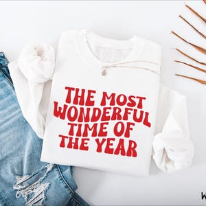 May include: A white long-sleeve sweatshirt featuring the red text THE MOST WONDERFUL TIME OF THE YEAR in a wavy font, paired with blue denim jeans. The sweatshirt has a crew neckline and a relaxed fit, showcasing a clean and simple design.