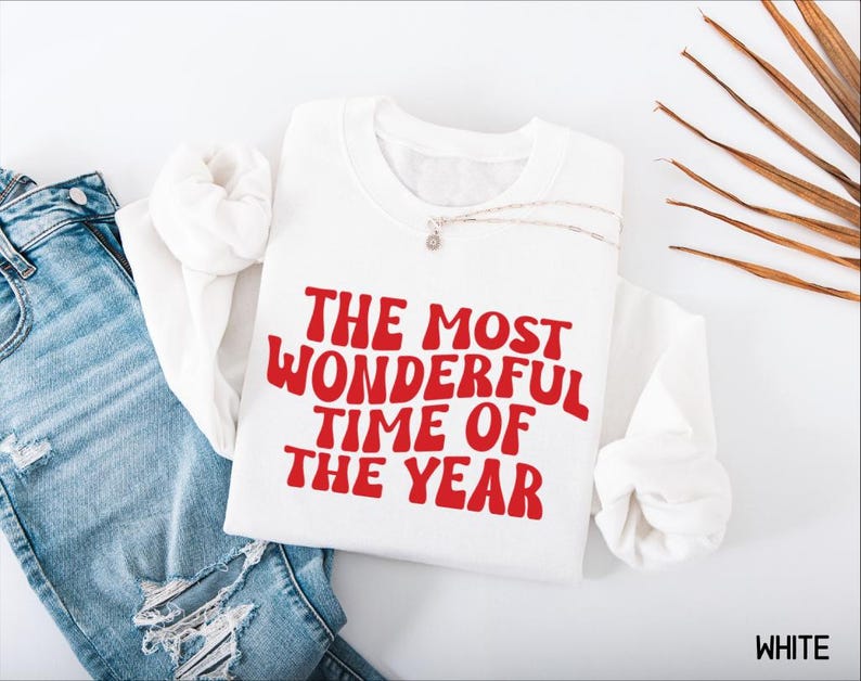 May include: A white long-sleeve sweatshirt featuring the red text THE MOST WONDERFUL TIME OF THE YEAR in a wavy font, paired with blue denim jeans. The sweatshirt has a crew neckline and a relaxed fit, showcasing a clean and simple design.