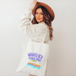 Puede incluir: Bolso tote color crema con la frase "Whatever Floats Your" en morado y un gr&aacute;fico colorido de un barco. La persona lleva una chaqueta color beige y un sombrero marr&oacute;n.