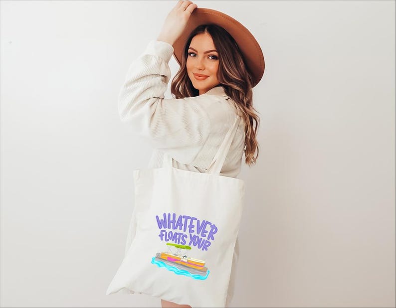 Puede incluir: Bolso tote color crema con la frase "Whatever Floats Your" en morado y un gr&aacute;fico colorido de un barco. La persona lleva una chaqueta color beige y un sombrero marr&oacute;n.