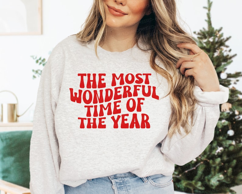 May include: A light gray crewneck sweatshirt featuring the red text "THE MOST WONDERFUL TIME OF THE YEAR" printed in a bold, wavy, retro-inspired font across the chest. The garment has long sleeves with ribbed cuffs and a ribbed neckline.