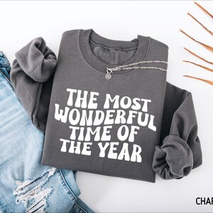 May include: A charcoal gray crewneck sweatshirt featuring the white text THE MOST WONDERFUL TIME OF THE YEAR in a wavy, retro-inspired font. The garment is displayed alongside a pair of light blue distressed denim jeans and a silver chain necklace.