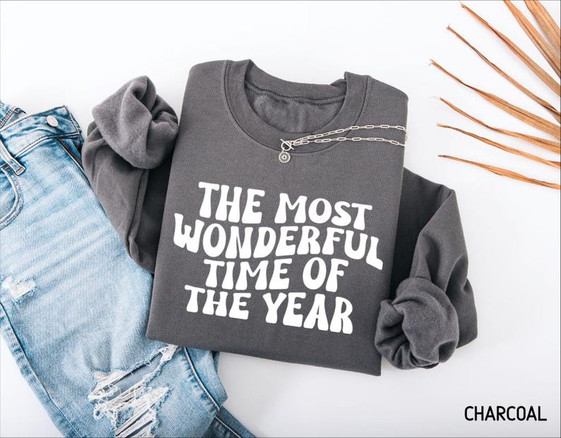 May include: A charcoal gray crewneck sweatshirt featuring the white text THE MOST WONDERFUL TIME OF THE YEAR in a wavy, retro-inspired font. The garment is displayed alongside a pair of light blue distressed denim jeans and a silver chain necklace.