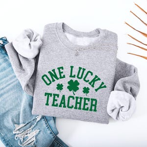 以下が含まれることがあります： 緑色の「ONE LUCKY TEACHER」の文字と3つの緑色のクローバーがプリントされた、ヘザーグレーのスウェットシャツ。スウェットシャツは青いジーンズと組み合わせています。シルバーのネックレスも見えます。