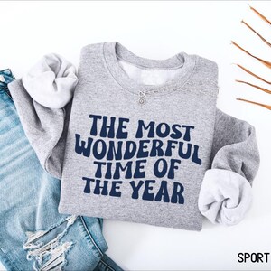 May include: A sport grey crewneck sweatshirt featuring the dark blue text THE MOST WONDERFUL TIME OF THE YEAR in a wavy font. The sweatshirt is displayed alongside a pair of distressed blue denim jeans and a silver chain necklace.