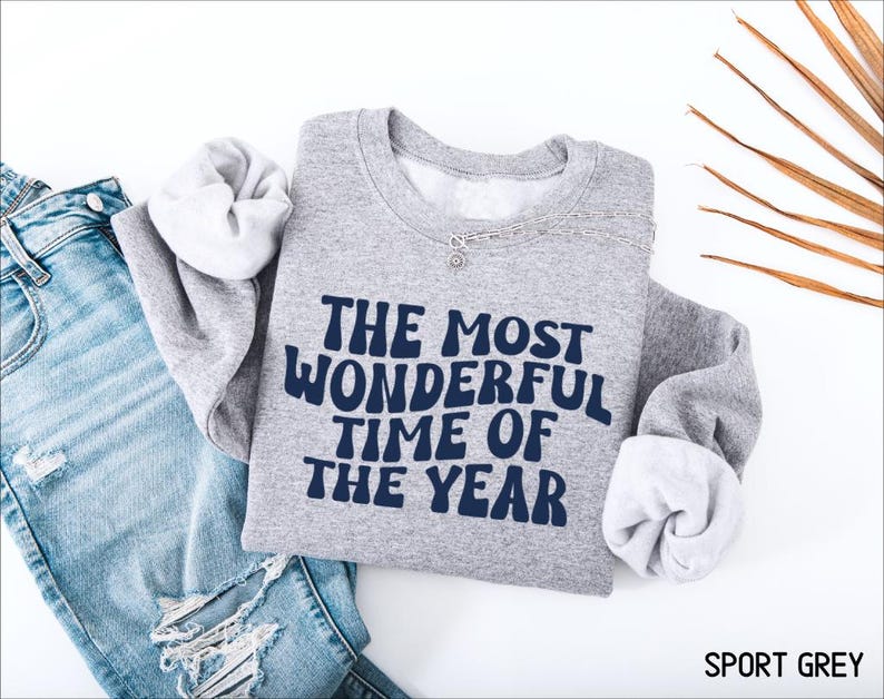 May include: A sport grey crewneck sweatshirt featuring the dark blue text THE MOST WONDERFUL TIME OF THE YEAR in a wavy font. The sweatshirt is displayed alongside a pair of distressed blue denim jeans and a silver chain necklace.
