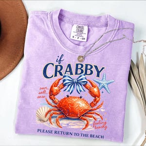 May include: A lilac Comfort Colors t-shirt with a crab graphic and the text "if CRABBY, PLEASE RETURN TO THE BEACH." The design includes a crab, bow, starfish, and seashell. A silver necklace and a brown hat are also visible.