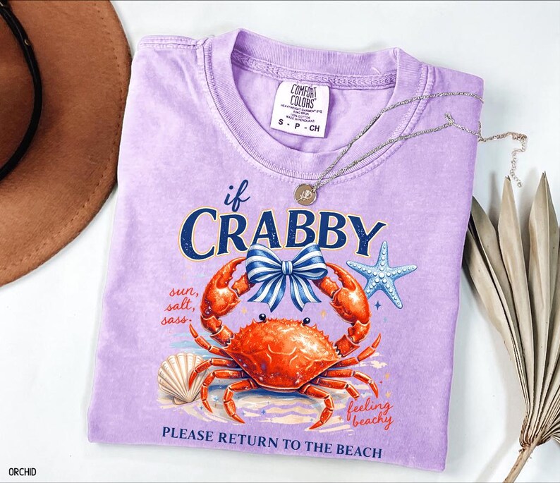 May include: A lilac Comfort Colors t-shirt with a crab graphic and the text "if CRABBY, PLEASE RETURN TO THE BEACH." The design includes a crab, bow, starfish, and seashell. A silver necklace and a brown hat are also visible.