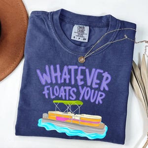 Puede incluir: Camiseta azul marino con la frase "WHATEVER FLOATS YOUR" en letras moradas. Una ilustraci&oacute;n colorida de un barco pont&oacute;n est&aacute; debajo del texto. La camiseta tiene cuello redondo y una peque&ntilde;a etiqueta.