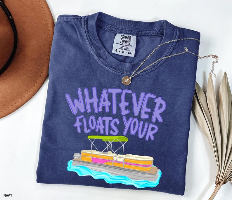 Puede incluir: Camiseta azul marino con la frase "WHATEVER FLOATS YOUR" en letras moradas. Una ilustraci&oacute;n colorida de un barco pont&oacute;n est&aacute; debajo del texto. La camiseta tiene cuello redondo y una peque&ntilde;a etiqueta.