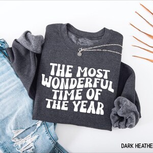 May include: A dark heather grey crewneck sweatshirt featuring the white text THE MOST WONDERFUL TIME OF THE YEAR in a bold, wavy font. The garment has long sleeves and a ribbed neckline, paired with light blue distressed denim jeans.
