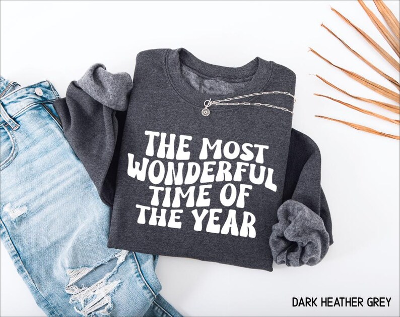 May include: A dark heather grey crewneck sweatshirt featuring the white text THE MOST WONDERFUL TIME OF THE YEAR in a bold, wavy font. The garment has long sleeves and a ribbed neckline, paired with light blue distressed denim jeans.