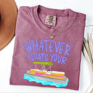 Puede incluir: Camiseta color bayas con la frase "WHATEVER FLOATS YOUR" en letras moradas. Un dibujo animado de un pont&oacute;n con toldo verde y detalles rosas y amarillos est&aacute; debajo del texto. Un collar dorado est&aacute; sobre la camiseta.