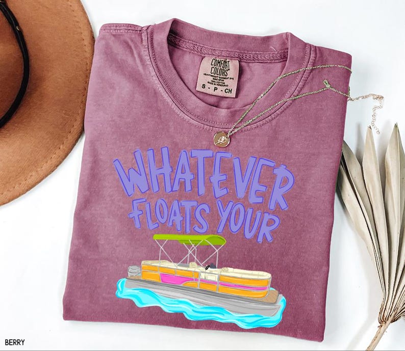 Puede incluir: Camiseta color bayas con la frase "WHATEVER FLOATS YOUR" en letras moradas. Un dibujo animado de un pont&oacute;n con toldo verde y detalles rosas y amarillos est&aacute; debajo del texto. Un collar dorado est&aacute; sobre la camiseta.
