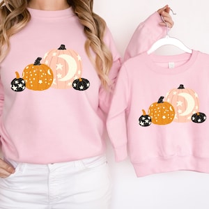 May include: Two pink sweatshirts with a graphic of three orange pumpkins with black stars and a crescent moon. The pumpkins are arranged in a row with the largest pumpkin on the right.