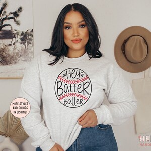 May include: Light gray sweatshirt with a baseball graphic that says "Hey Batter Batter" in black and red. The sweatshirt has long sleeves and a crew neck. The image also includes text that says "More styles and colors in listing!" and "Hello Handpressed".
