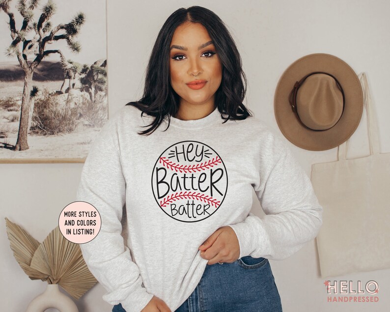 May include: Light gray sweatshirt with a baseball graphic that says "Hey Batter Batter" in black and red. The sweatshirt has long sleeves and a crew neck. The image also includes text that says "More styles and colors in listing!" and "Hello Handpressed".