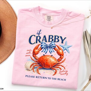 May include: A light pink t-shirt with a crab graphic. The crab is orange with a blue bow, and the text reads "if CRABBY, PLEASE RETURN TO THE BEACH." Additional text includes "sun, salt, sass" and "feeling beachy."