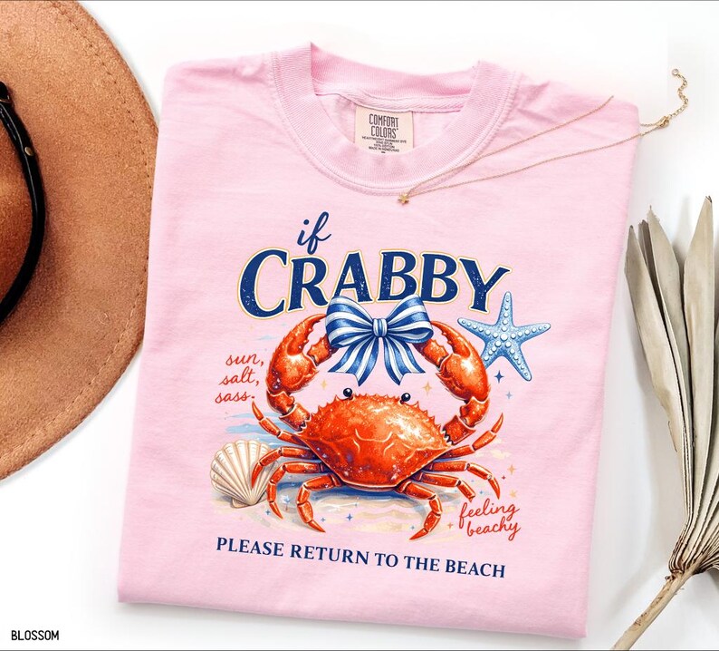 May include: A light pink t-shirt with a crab graphic. The crab is orange with a blue bow, and the text reads "if CRABBY, PLEASE RETURN TO THE BEACH." Additional text includes "sun, salt, sass" and "feeling beachy."
