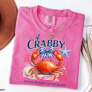 May include: A pink Comfort Colors t-shirt with a crab graphic and the text "if Crabby, Please Return to the Beach." The design includes a crab, bow, starfish, and seashell. A gold necklace is draped across the shirt.