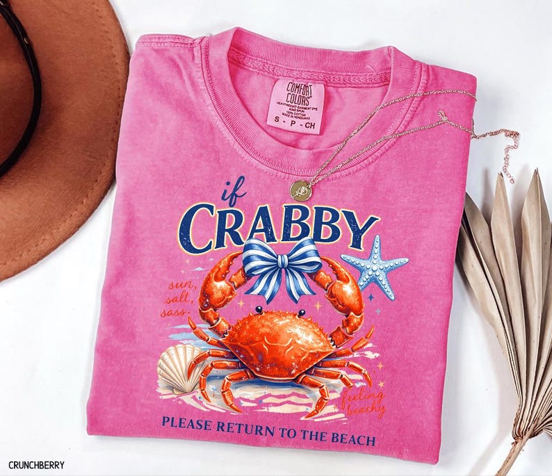 May include: A pink Comfort Colors t-shirt with a crab graphic and the text "if Crabby, Please Return to the Beach." The design includes a crab, bow, starfish, and seashell. A gold necklace is draped across the shirt.