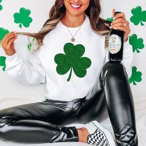 K&ouml;nnte beinhalten: Ein wei&szlig;es Sweatshirt mit einem gr&uuml;nen Glitzer-Kleeblatt-Design. Das Model tr&auml;gt eine schwarze Lederhose und h&auml;lt eine Flasche Guinness.