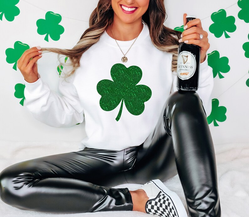 K&ouml;nnte beinhalten: Ein wei&szlig;es Sweatshirt mit einem gr&uuml;nen Glitzer-Kleeblatt-Design. Das Model tr&auml;gt eine schwarze Lederhose und h&auml;lt eine Flasche Guinness.