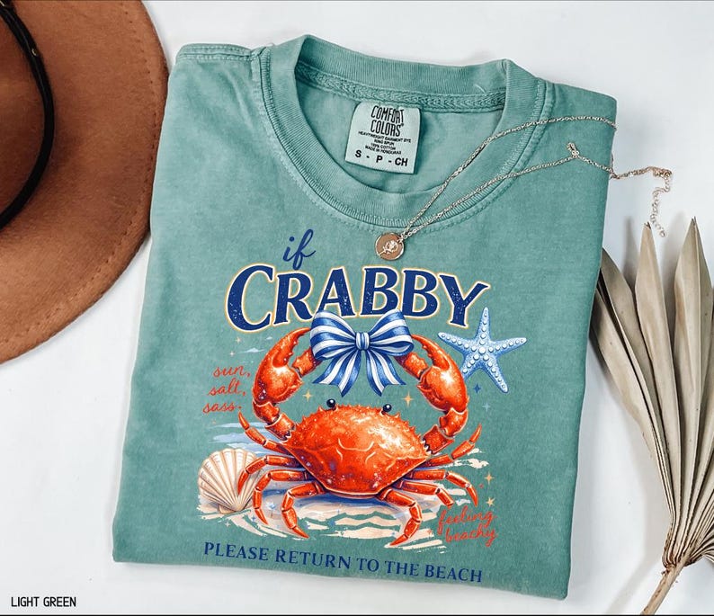 May include: Light green t-shirt with a graphic of a crab, bow, and starfish. The text on the shirt reads "if CRABBY, PLEASE RETURN TO THE BEACH". A silver necklace with a pendant is on the shirt.