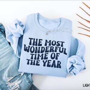 May include: A light blue crewneck sweatshirt featuring the dark blue text THE MOST WONDERFUL TIME OF THE YEAR in a wavy font, paired with distressed blue denim jeans and a silver chain necklace resting on the collar of the sweatshirt.