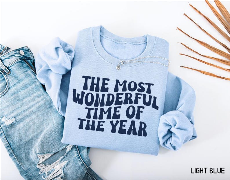 May include: A light blue crewneck sweatshirt featuring the dark blue text THE MOST WONDERFUL TIME OF THE YEAR in a wavy font, paired with distressed blue denim jeans and a silver chain necklace resting on the collar of the sweatshirt.