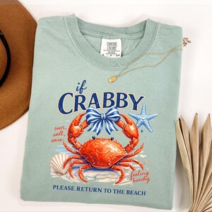 May include: A light green t-shirt with a crab graphic and the text "if CRABBY, PLEASE RETURN TO THE BEACH". The graphic includes a red crab, blue bow, starfish, and seashell. A gold necklace is draped across the shirt.