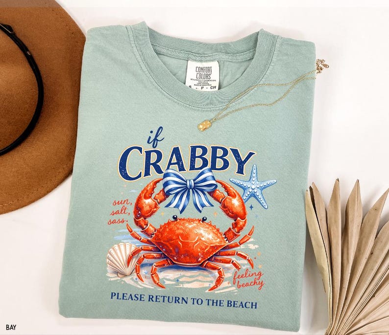 May include: A light green t-shirt with a crab graphic and the text "if CRABBY, PLEASE RETURN TO THE BEACH". The graphic includes a red crab, blue bow, starfish, and seashell. A gold necklace is draped across the shirt.